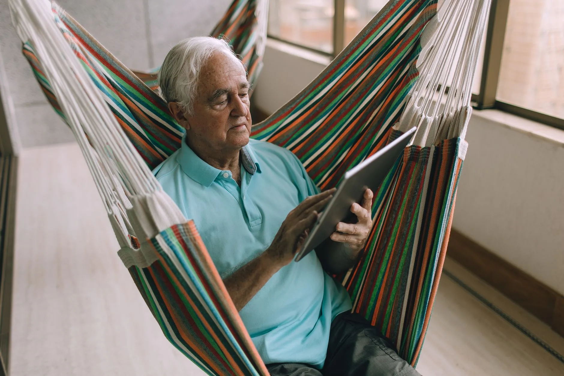 Elderly man using a tablet while sitting comfortably in a hammock indoors.
