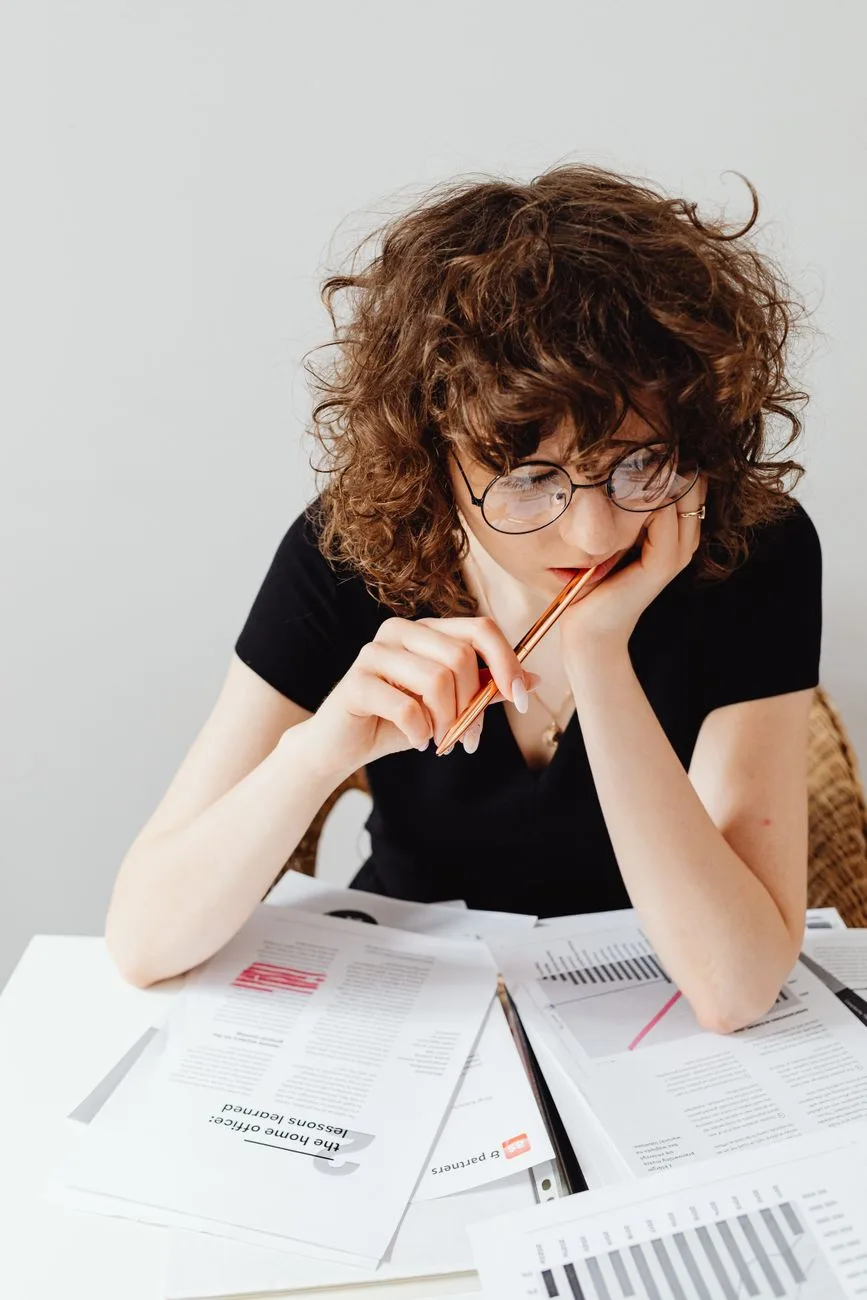 Curly-haired woman in glasses focuses on documents at her workspace, analyzing and working.