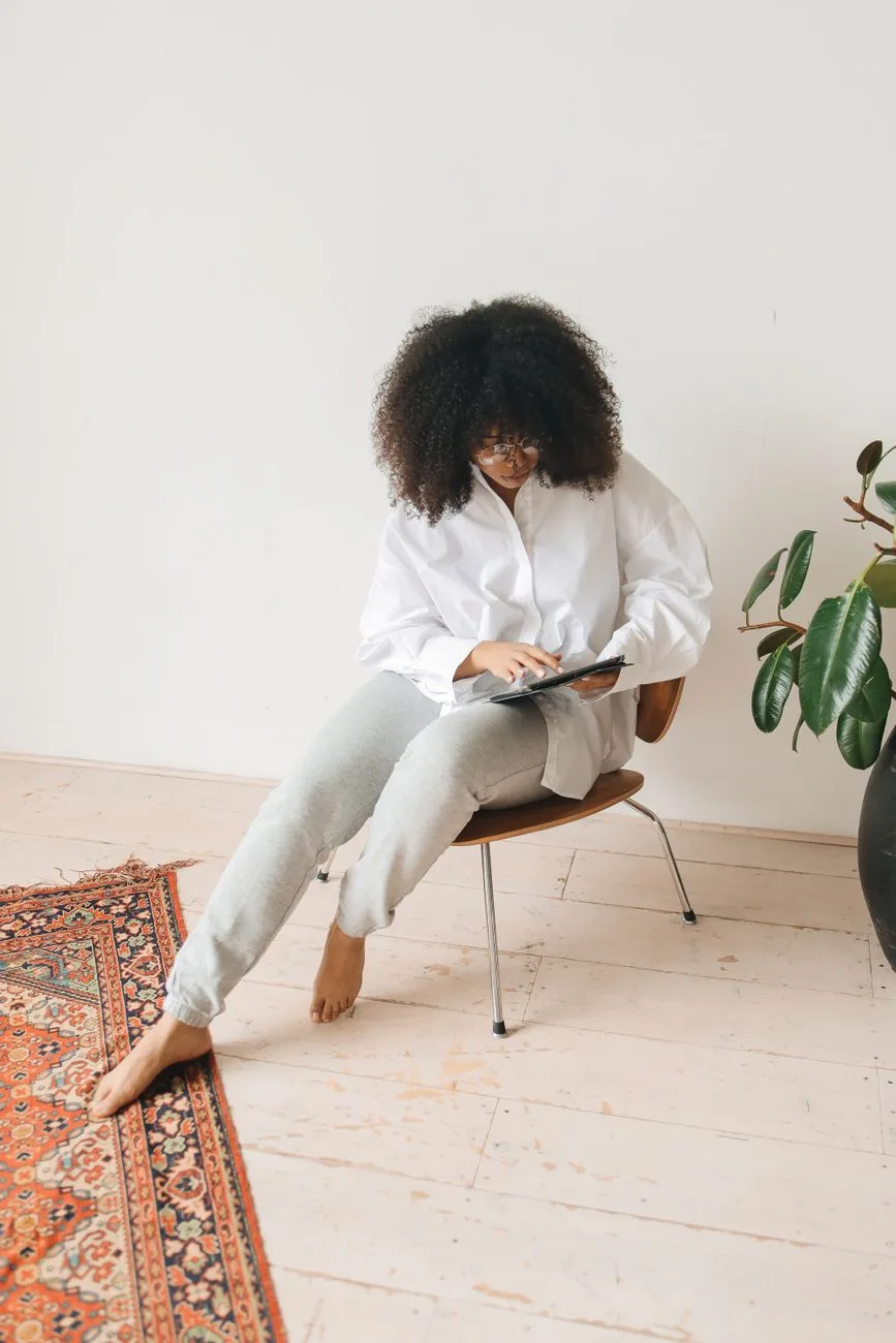 A woman with curly hair sits on a chair using a tablet, embodying a relaxed work-from-home setup.