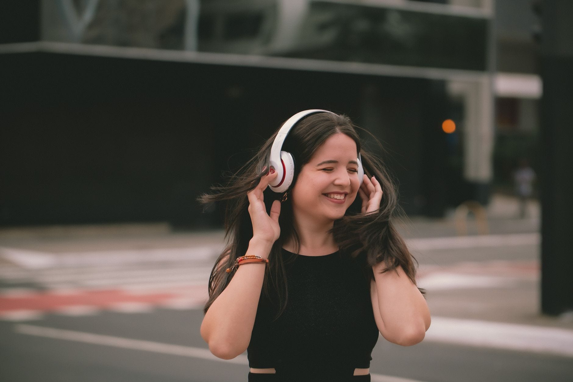 A smiling woman listens to music wearing wireless headphones in an urban setting, expressing happiness.